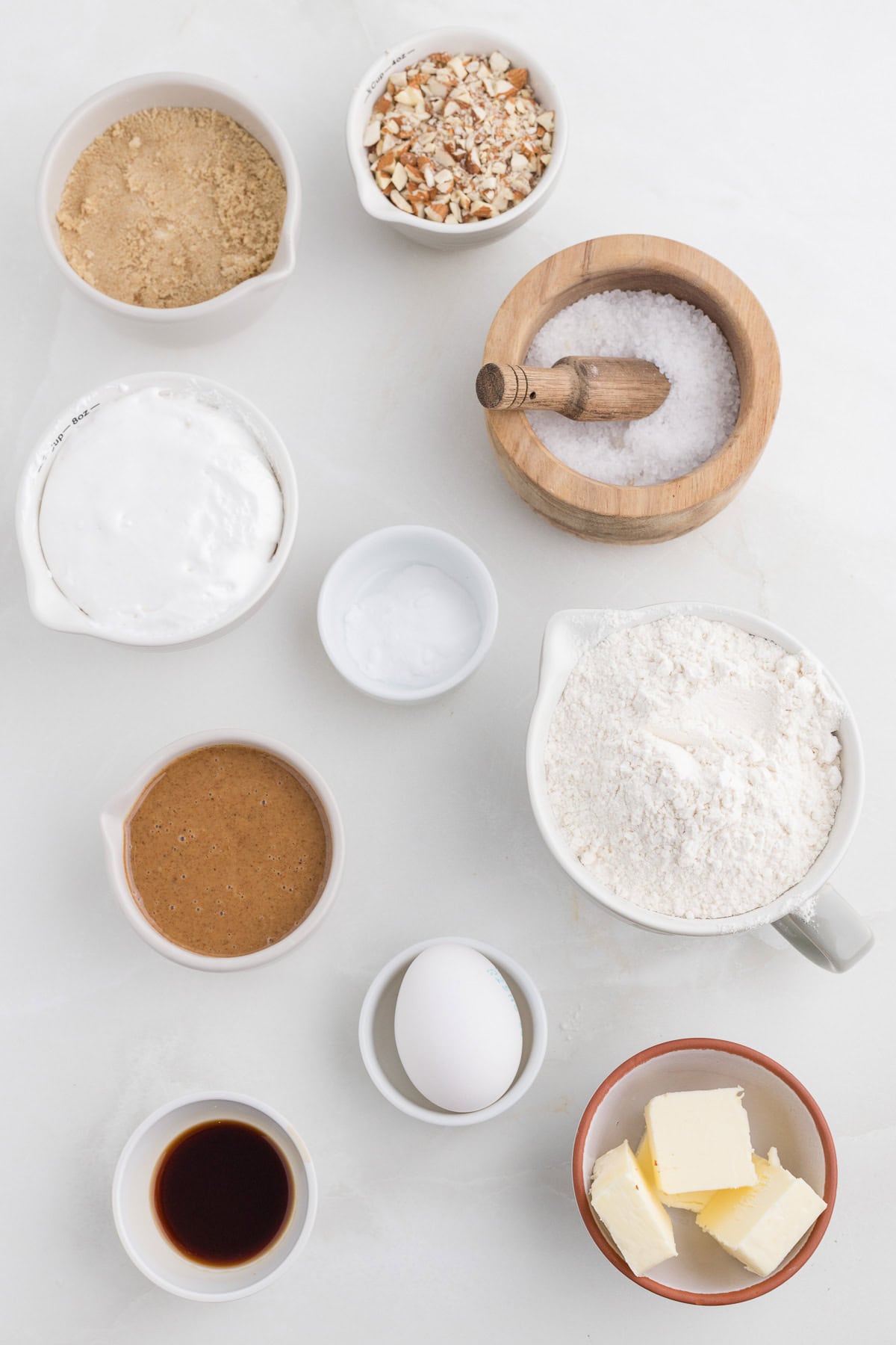 Assorted baking ingredients including flour, an egg, butter, salt, brown sugar, vanilla extract, coconut cream, chopped nuts, almond butter, and baking powder arranged on a white surface.
