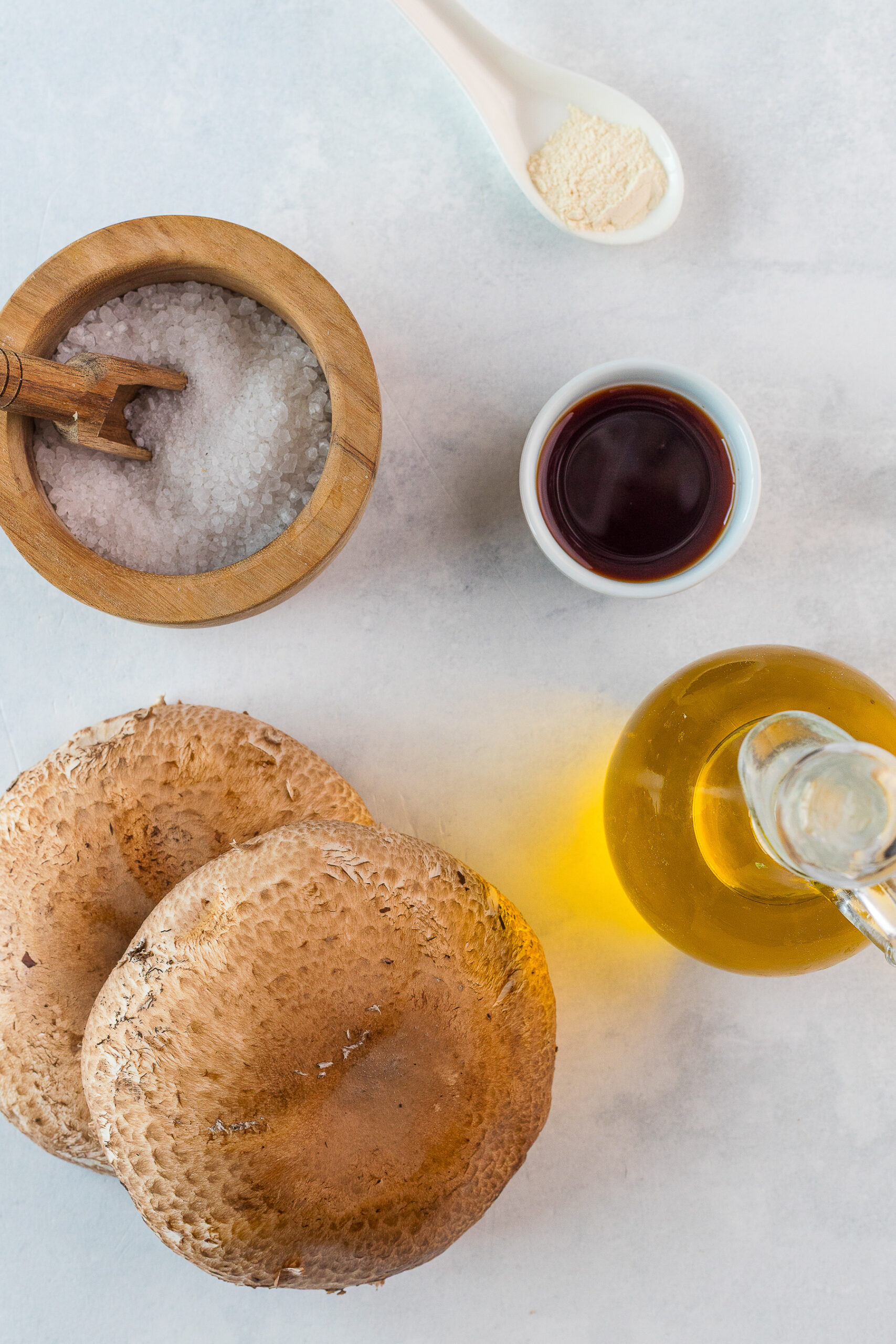 Two large portobello mushrooms, a wooden bowl of salt, a white spoon with garlic powder, a small dish of soy sauce, and a glass jar of olive oil on a light surface.