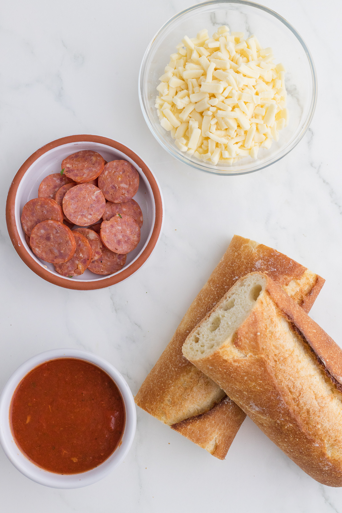 Sliced baguette, a bowl of shredded cheese, a bowl of sliced pepperoni, and a bowl of red sauce arranged on a white surface.