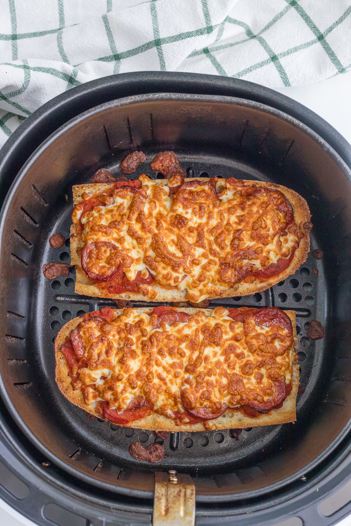 Two slices of French bread pizza with melted cheese and pepperoni inside an air fryer basket, with a checkered kitchen towel in the background.