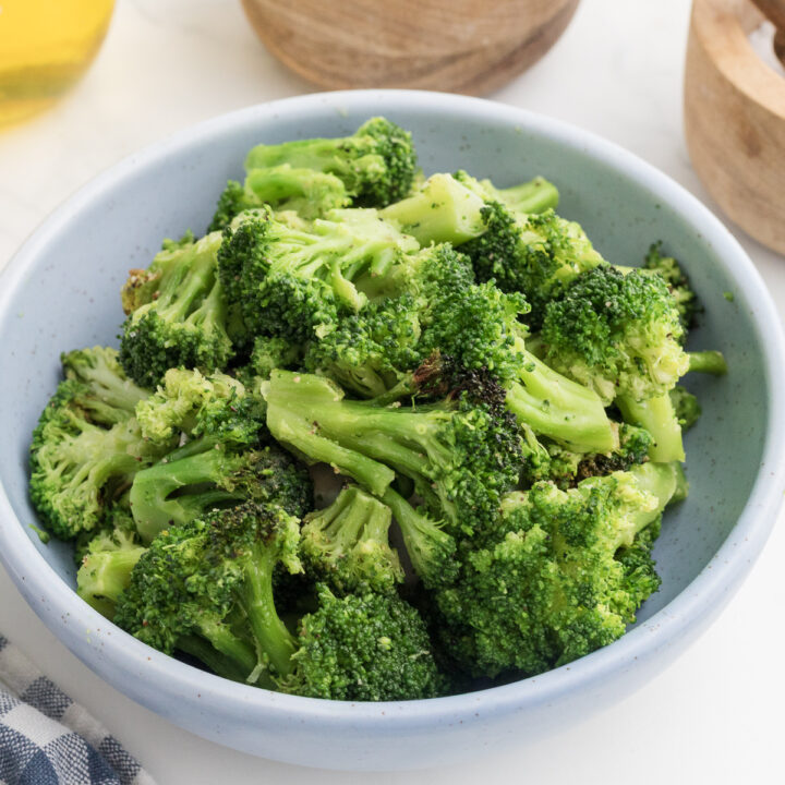 A bowl of cooked broccoli florets in a light blue dish, placed on a white surface with part of a napkin and wooden bowls visible nearby.