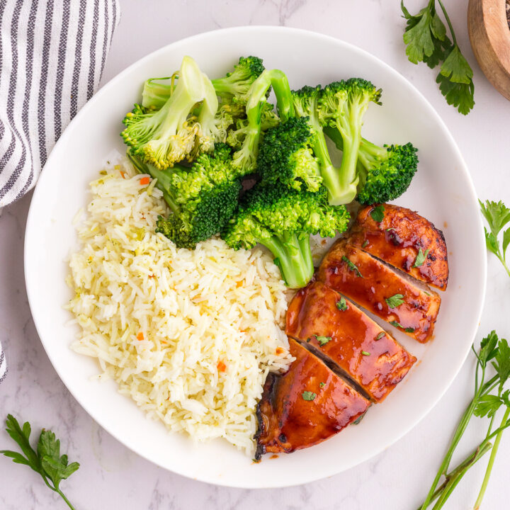 A white plate with steamed broccoli, seasoned white rice, and sliced glazed chicken breast on a marble surface.