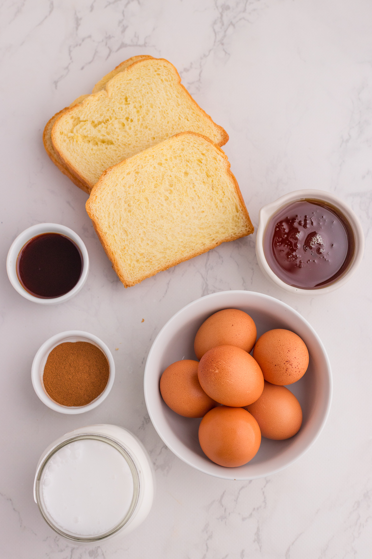Two slices of bread, six brown eggs in a bowl, a jar of milk, a bowl of cinnamon, a small cup of vanilla extract, and a bowl of jam on a white surface.