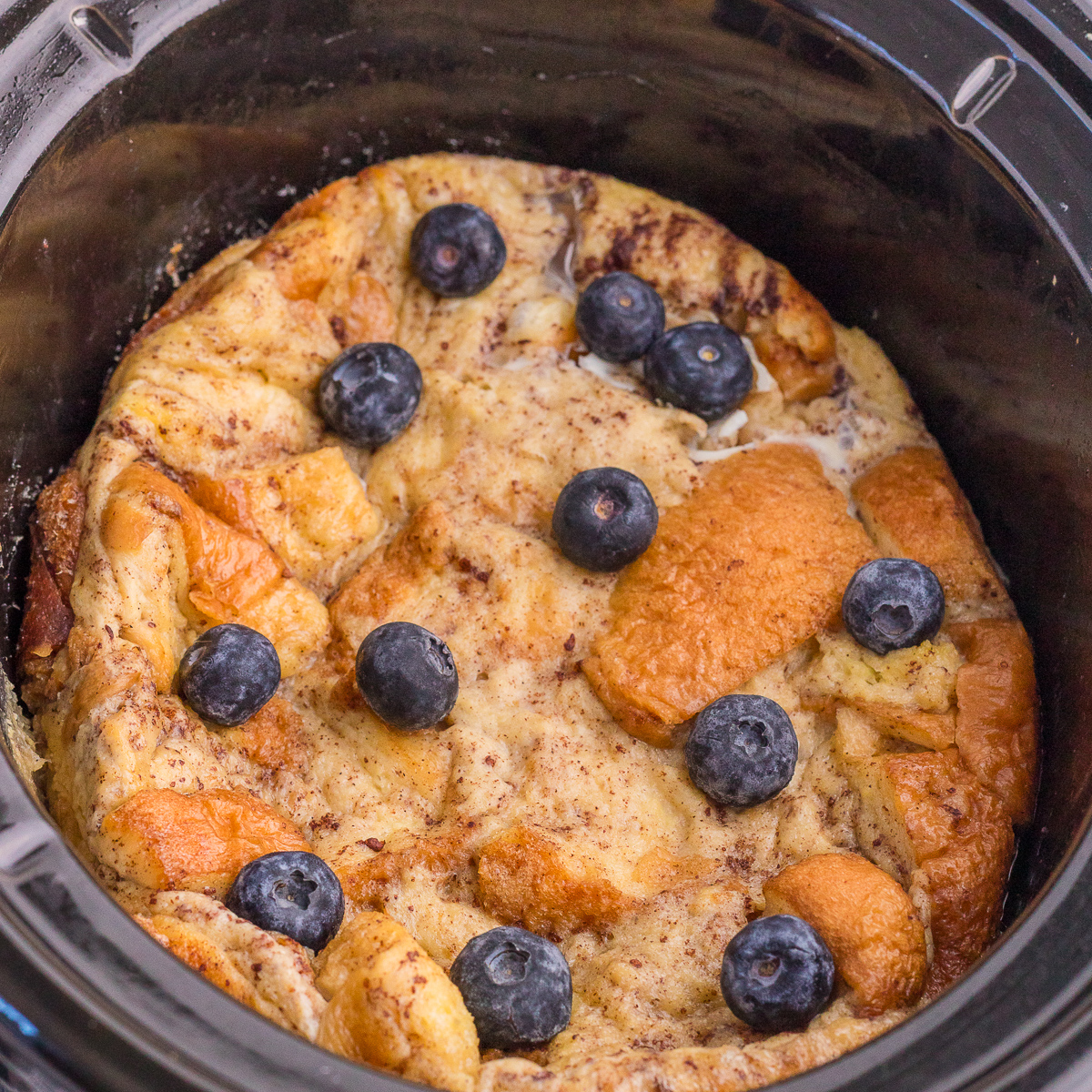 A baked bread pudding topped with blueberries is shown inside a slow cooker.