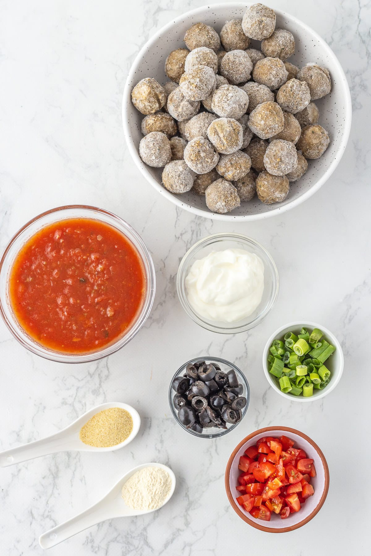 A bowl of frozen meatballs, enchilada sauce, sour cream, chopped green onions, sliced black olives, diced tomatoes, garlic powder, and onion powder on a white surface.