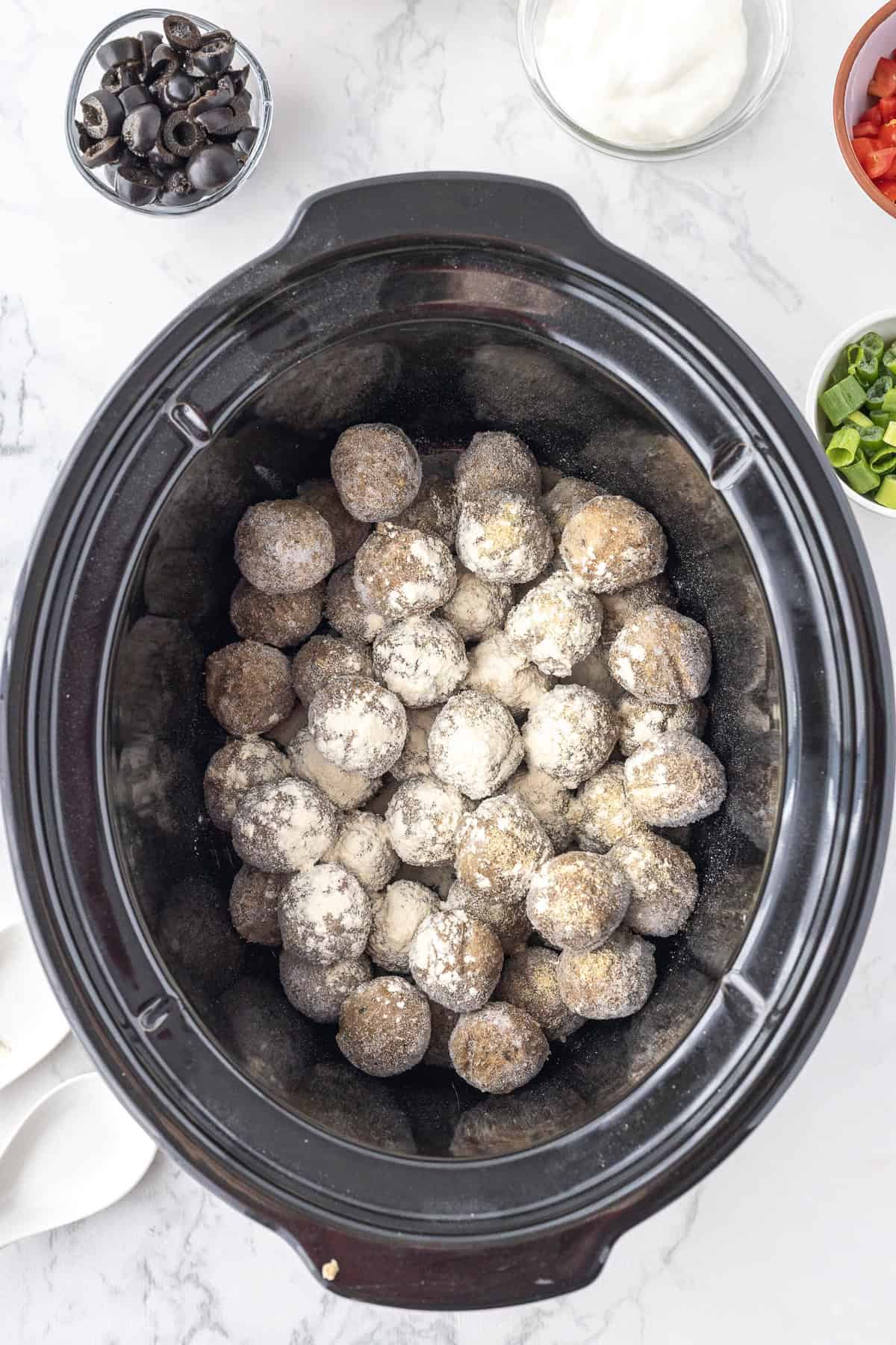 A slow cooker filled with uncooked,  meatballs, surrounded by small bowls of sliced olives, yogurt, chopped tomatoes, and green onions.
