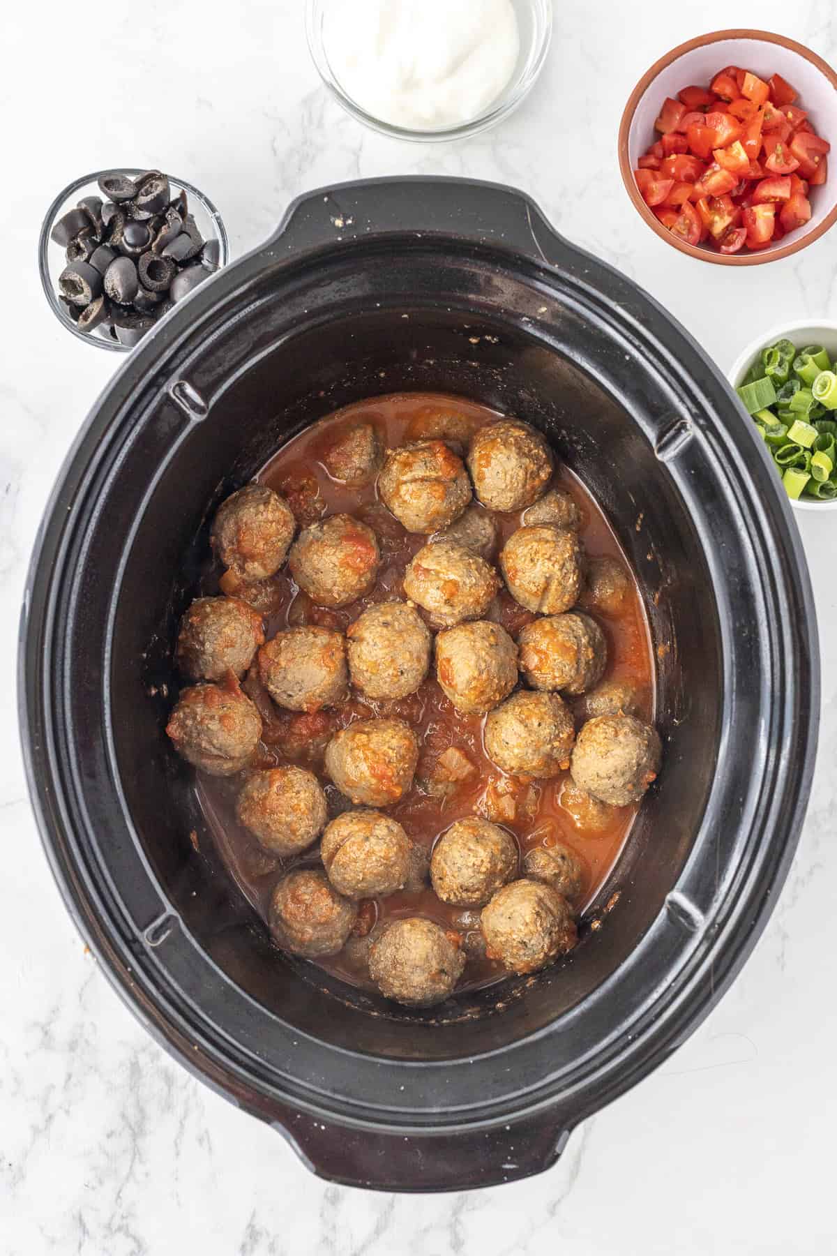 A slow cooker filled with meatballs in sauce, surrounded by small bowls of sliced black olives, chopped tomatoes, green onions, and a bowl of white sauce.