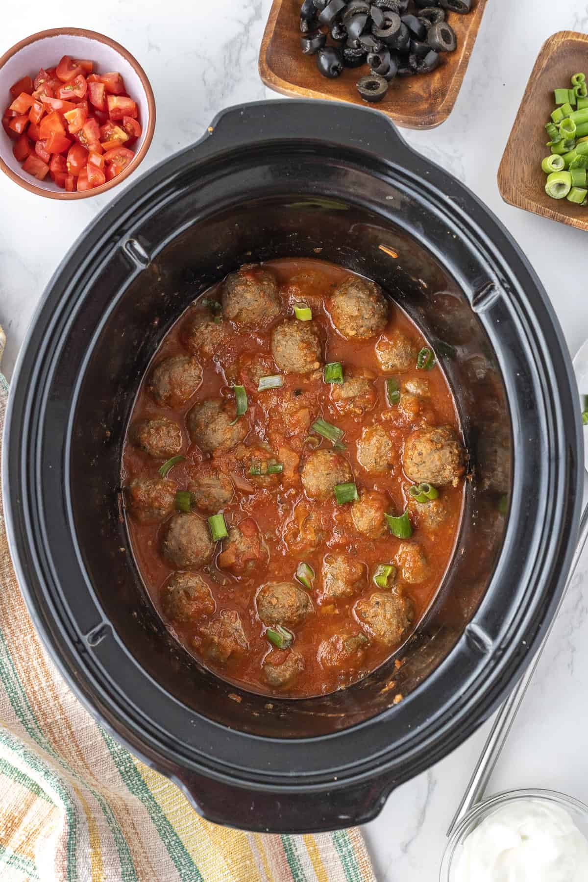 Meatballs in sauce garnished with chopped green onions in a slow cooker, surrounded by bowls of diced tomatoes, sliced olives, and green onions.