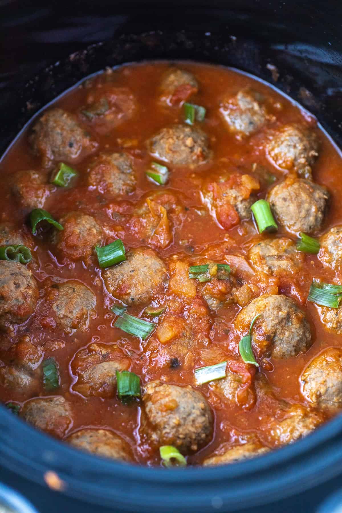 A slow cooker filled with meatballs in sauce, garnished with chopped green onions.