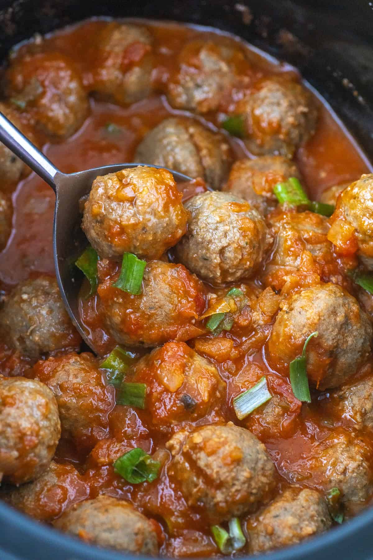 A ladle scoops meatballs in sauce from a pot, garnished with chopped green onions.