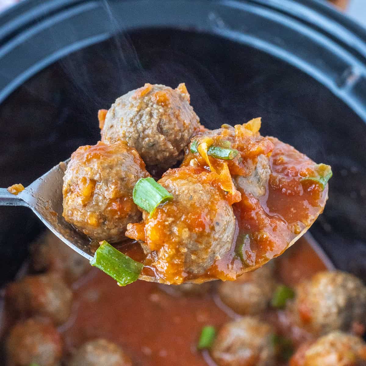 A spoon holds several meatballs covered in sauce with chopped green onions, above a slow cooker filled with more meatballs and sauce.