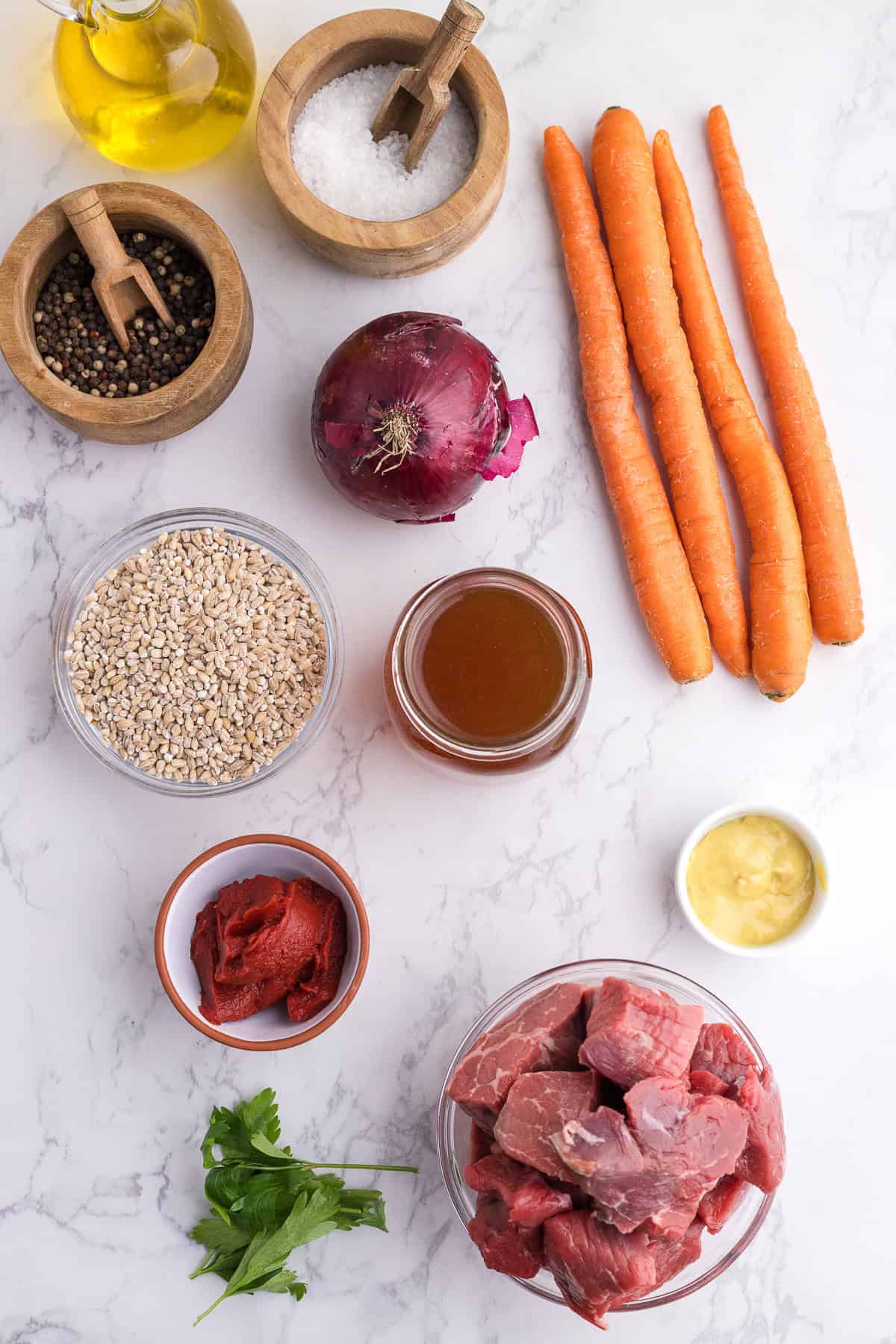 Top-down view of raw ingredients including carrots, beef cubes, barley, tomato paste, broth, onion, oil, parsley, minced garlic, salt, and pepper on a white marble surface.