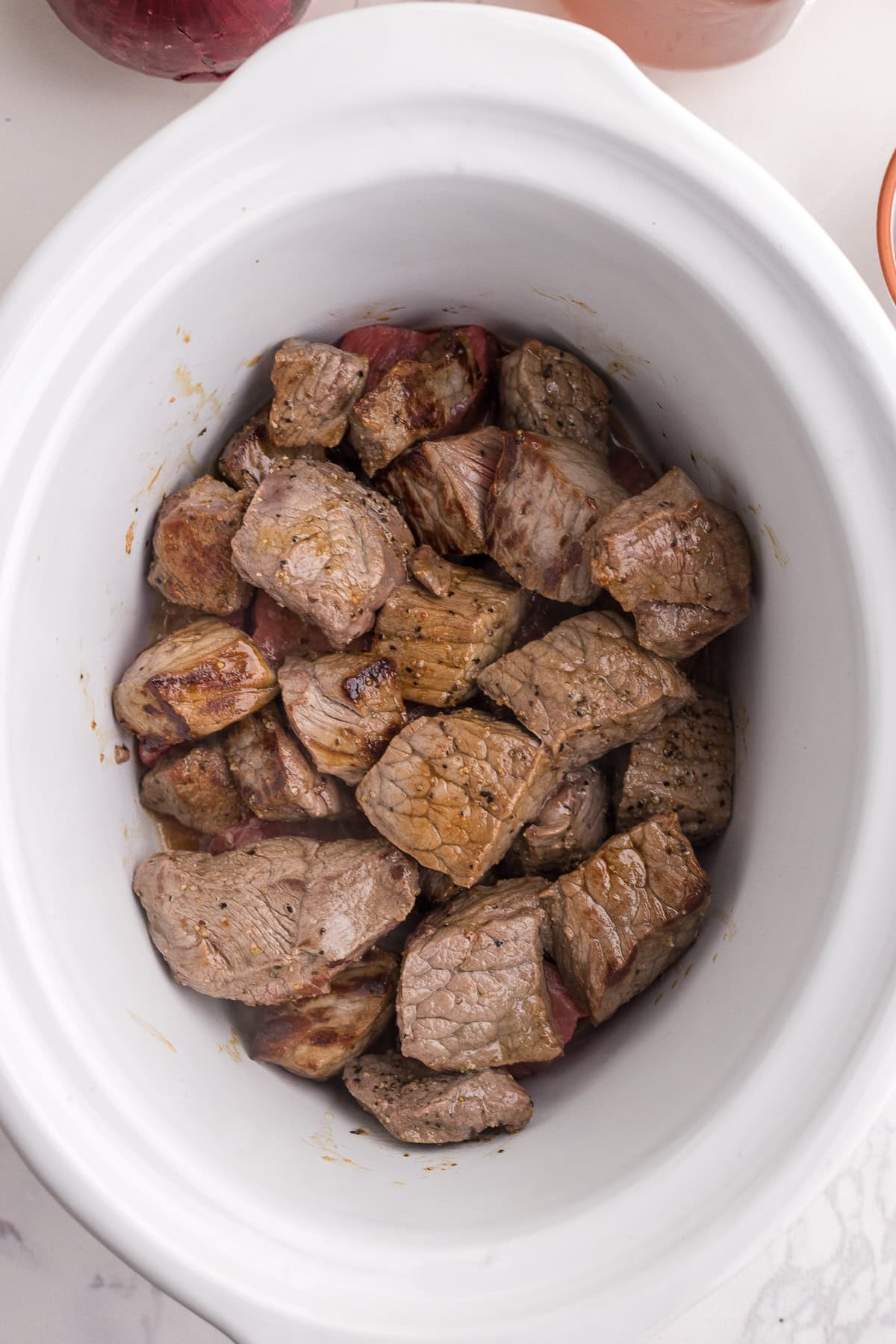 Chunks of browned beef are placed in a white slow cooker, ready for cooking.