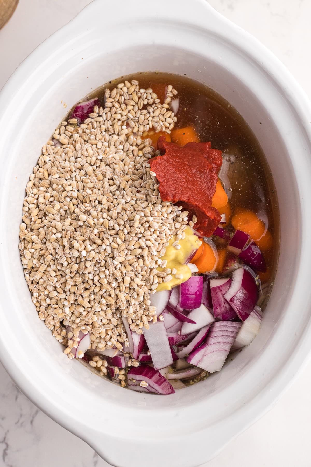 A slow cooker filled with uncooked barley, chopped red onion, carrot slices, garlic, tomato paste, and broth.