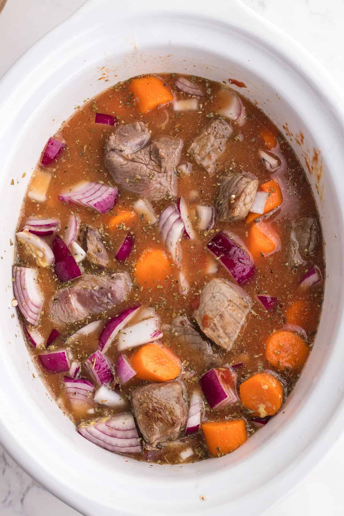 Chunks of beef, red onion, and carrot in seasoned broth inside a white slow cooker, ready for cooking.