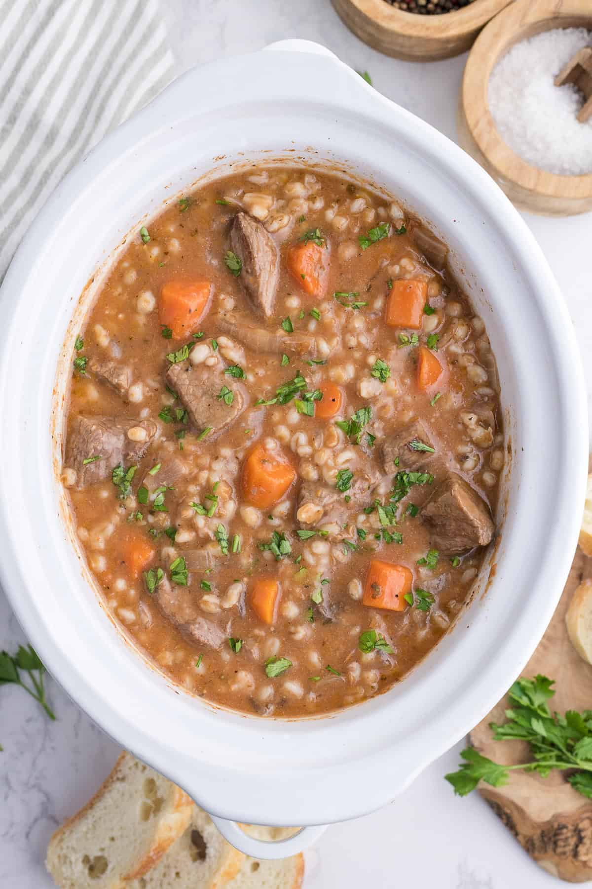 A white slow cooker filled with beef and barley stew, containing chunks of beef, carrots, and barley, garnished with chopped parsley.