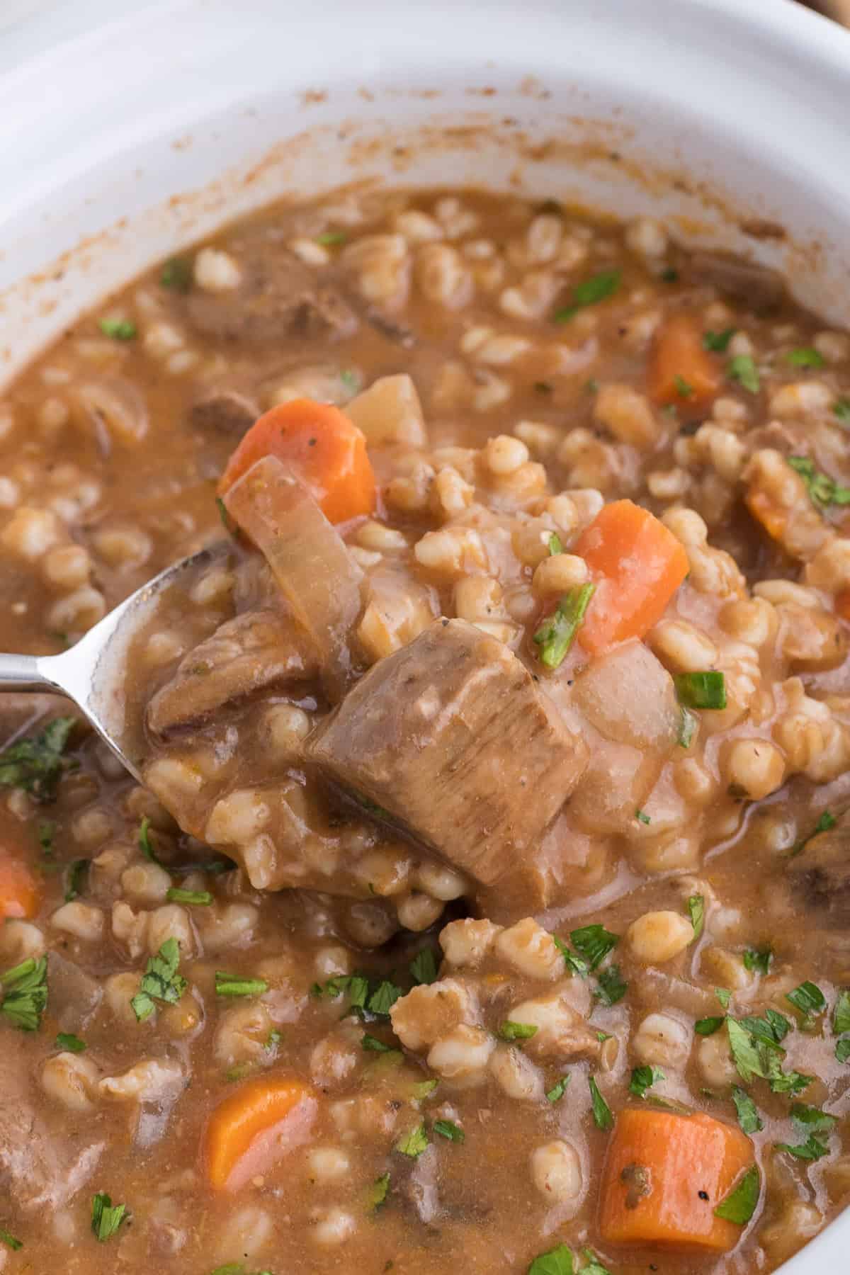 A close-up of beef and barley stew with carrots and parsley in a white bowl; a spoon lifts a portion of the chunky stew.