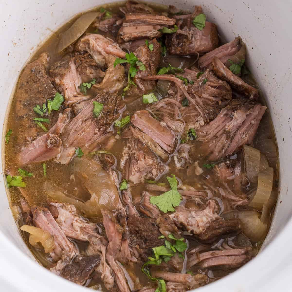 Shredded beef in broth with onions and chopped cilantro in a slow cooker.