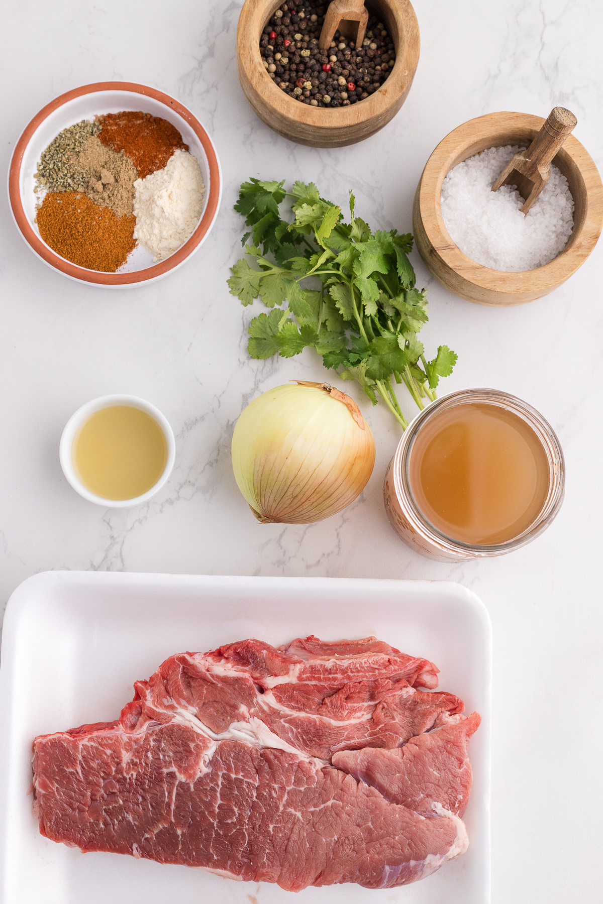 Raw beef steak, an onion, cilantro, spices, black peppercorns, salt, lime juice, and beef broth arranged on a white surface.