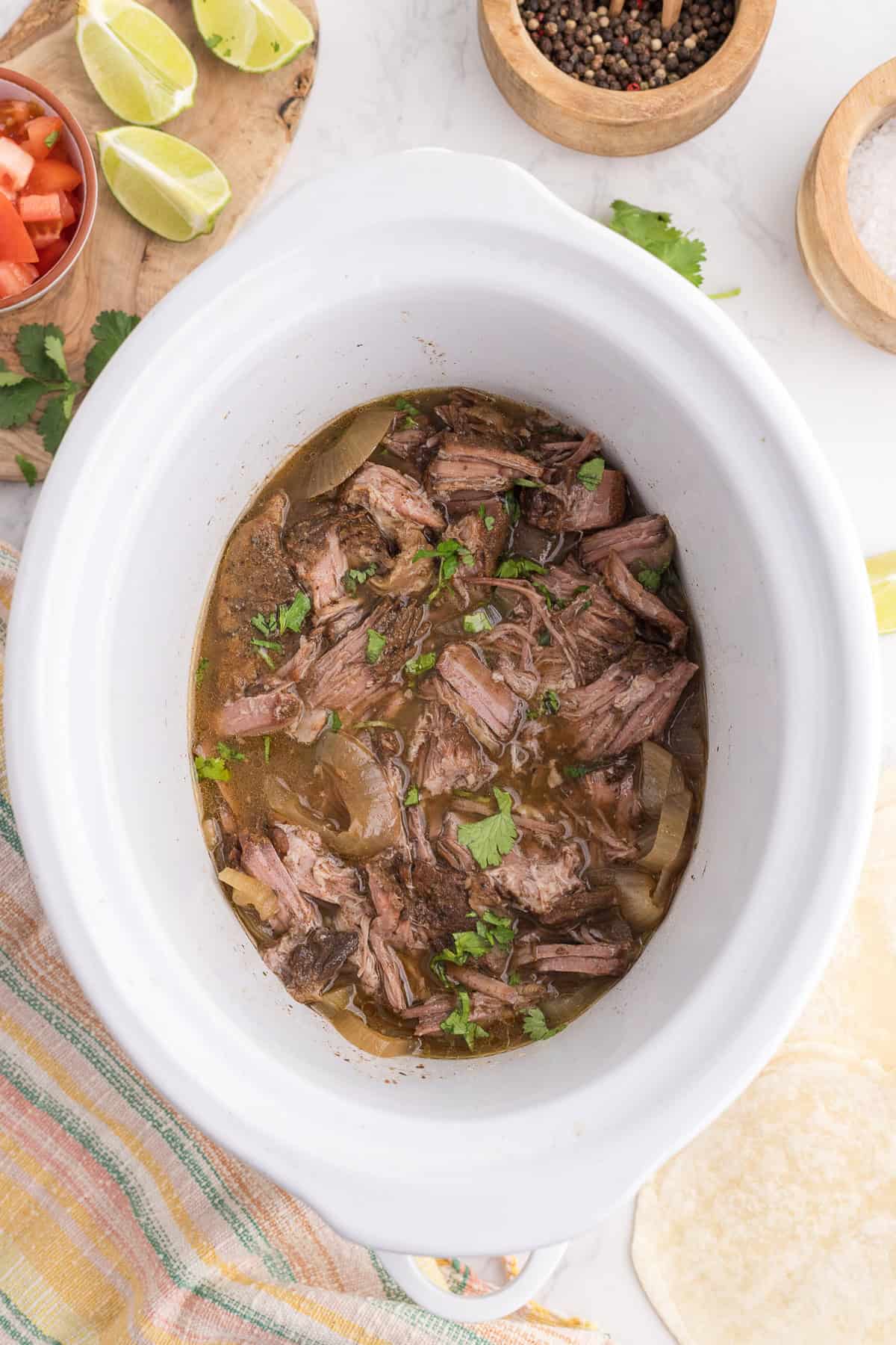 Shredded beef with herbs and broth in a white slow cooker, surrounded by lime wedges, chopped tomatoes, tortillas, and spices on a white countertop.