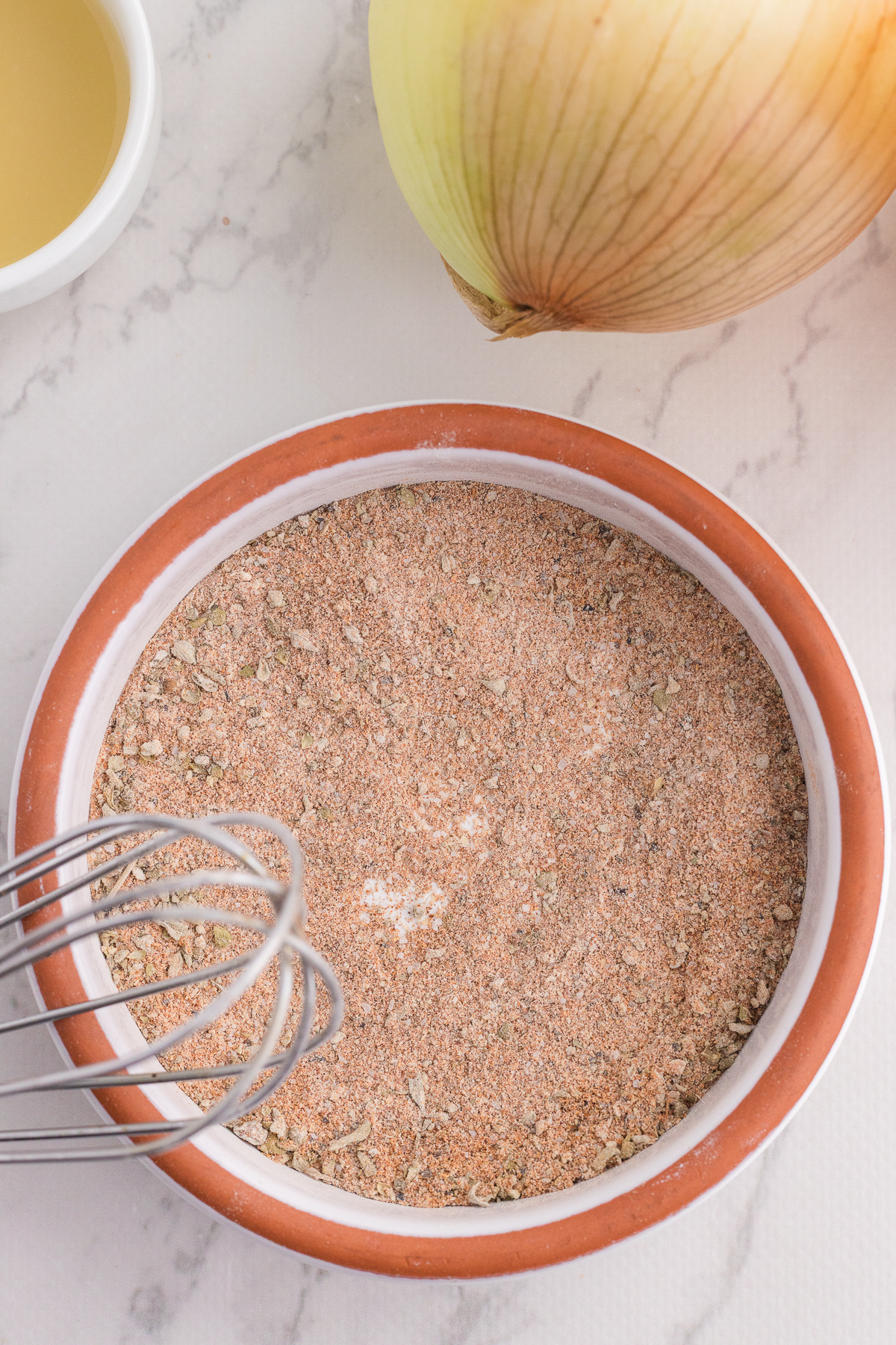 A ceramic bowl filled with a dry seasoning mix sits on a marble surface next to a whisk, a whole onion, and a small bowl of liquid.