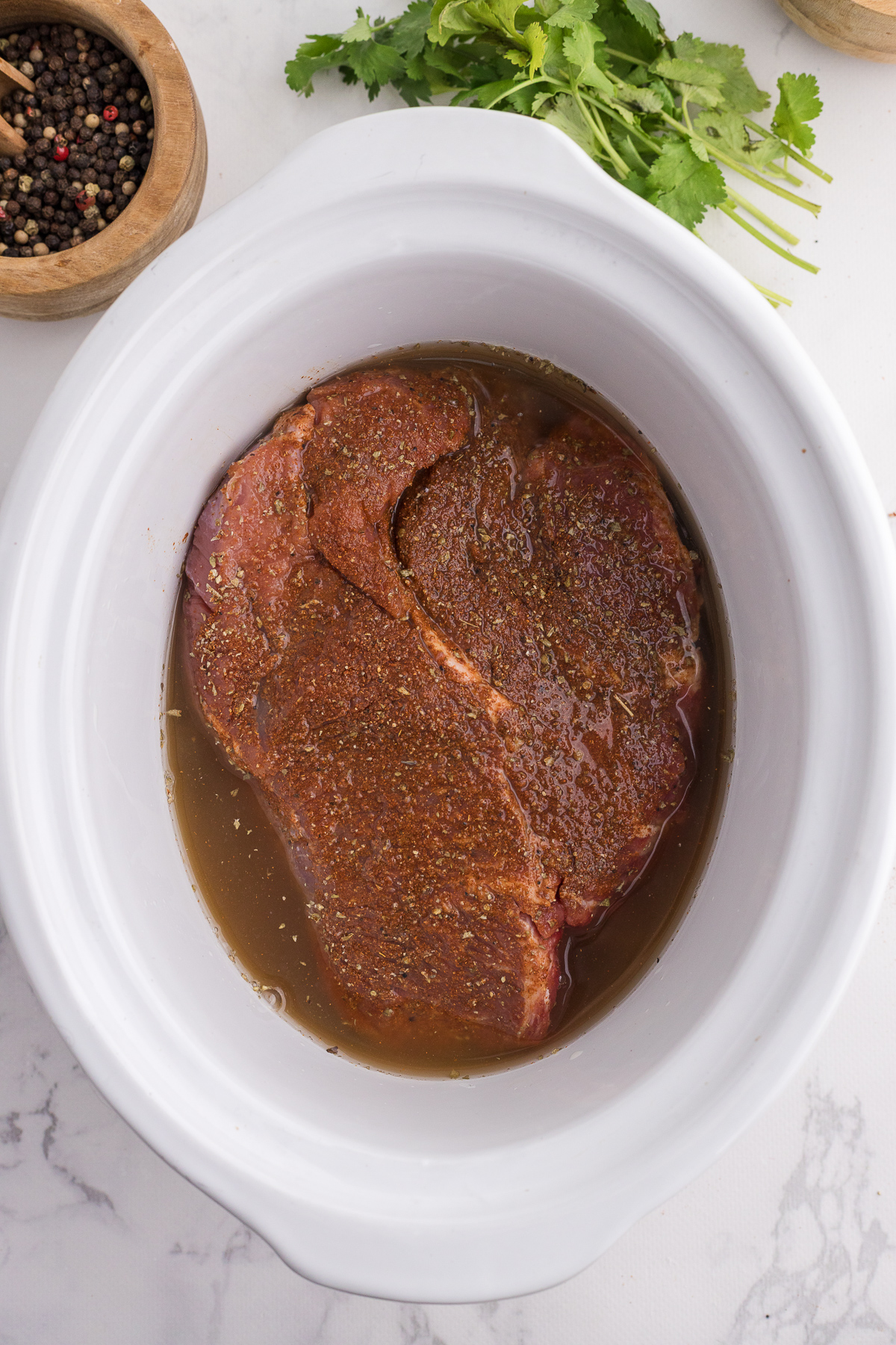 A seasoned piece of raw beef sits in broth inside a white slow cooker, with peppercorns and fresh cilantro nearby.