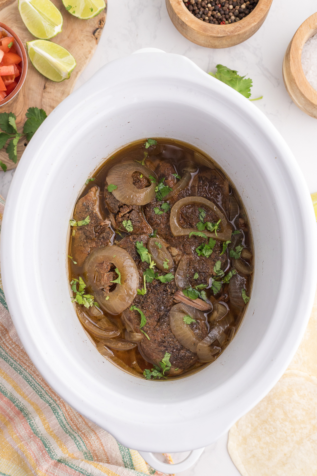 Slow cooker filled with cooked beef topped with sliced onions and chopped cilantro, surrounded by lime wedges, spices, and tortillas on a white surface.