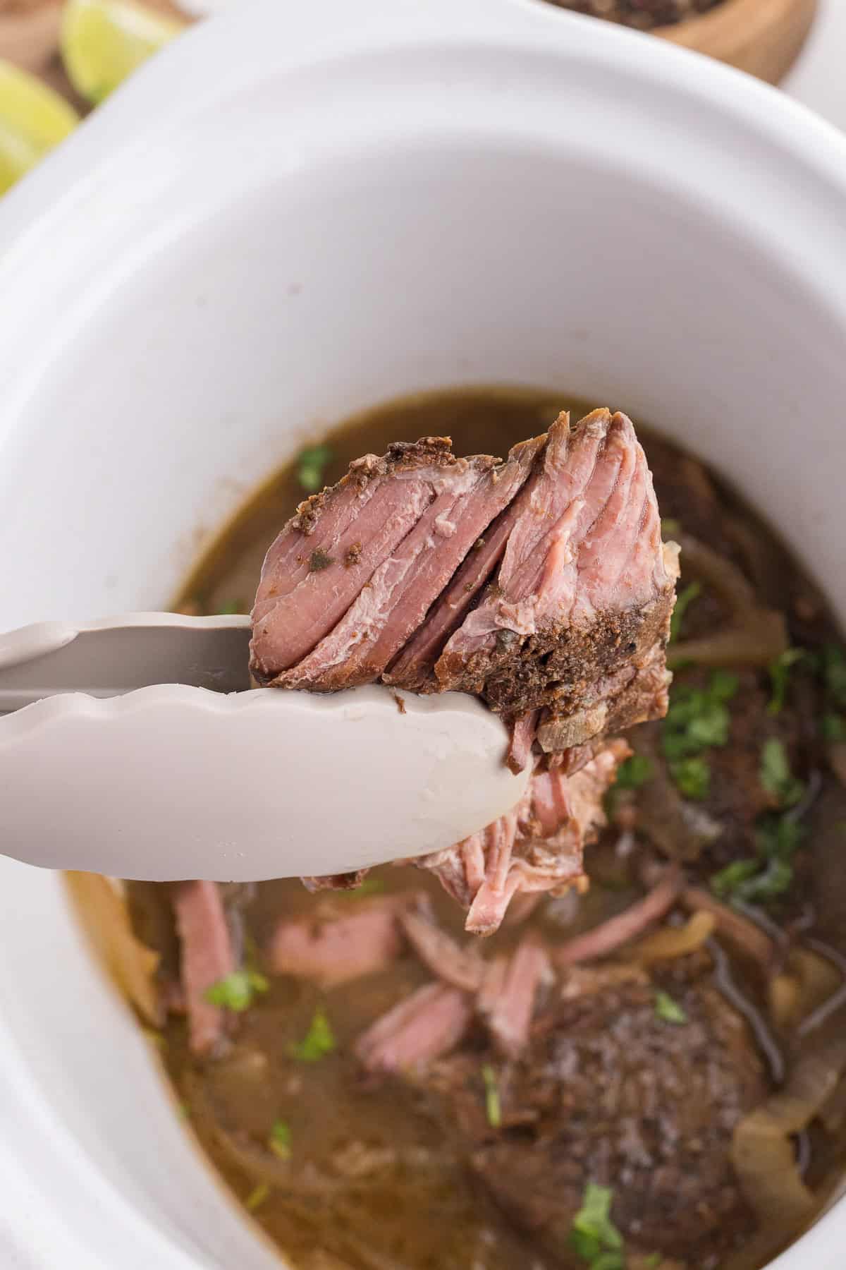 Metal tongs hold a piece of cooked, shredded beef above a white slow cooker filled with broth and herbs.