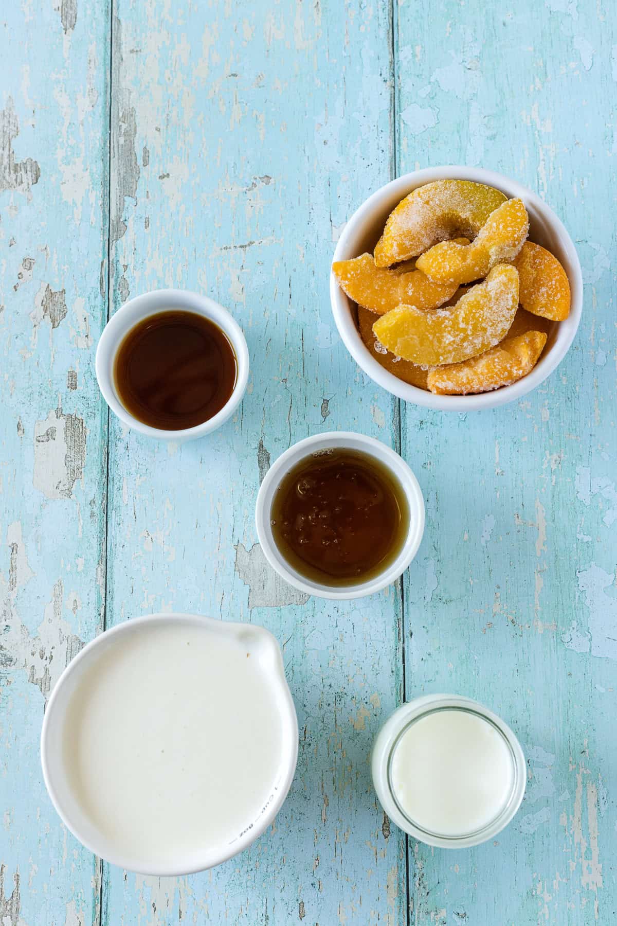 Five bowls containing dried peaches, two types of syrup, heavy cream, and milk are arranged on a light blue wooden surface—perfect ingredients for making Ninja Creami peach ice cream.