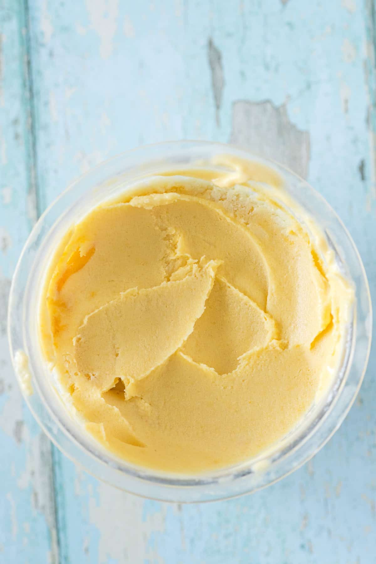 A clear container filled with smooth, light orange Ninja Creami peach ice cream sits on a distressed blue wooden surface.