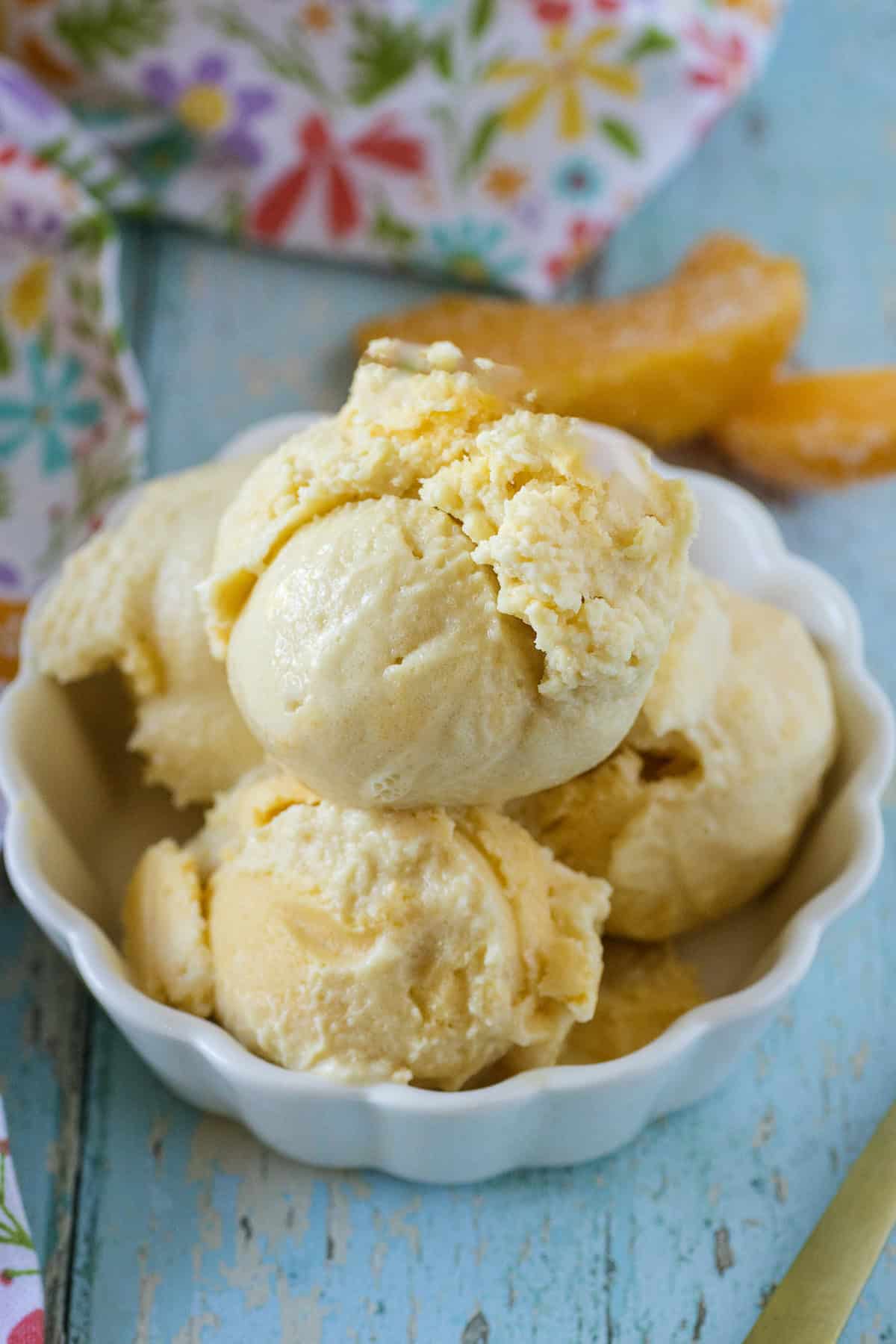 Three scoops of Ninja Creami peach ice cream sit in a small white bowl on a blue wooden surface, with a floral cloth and orange slices in the background.