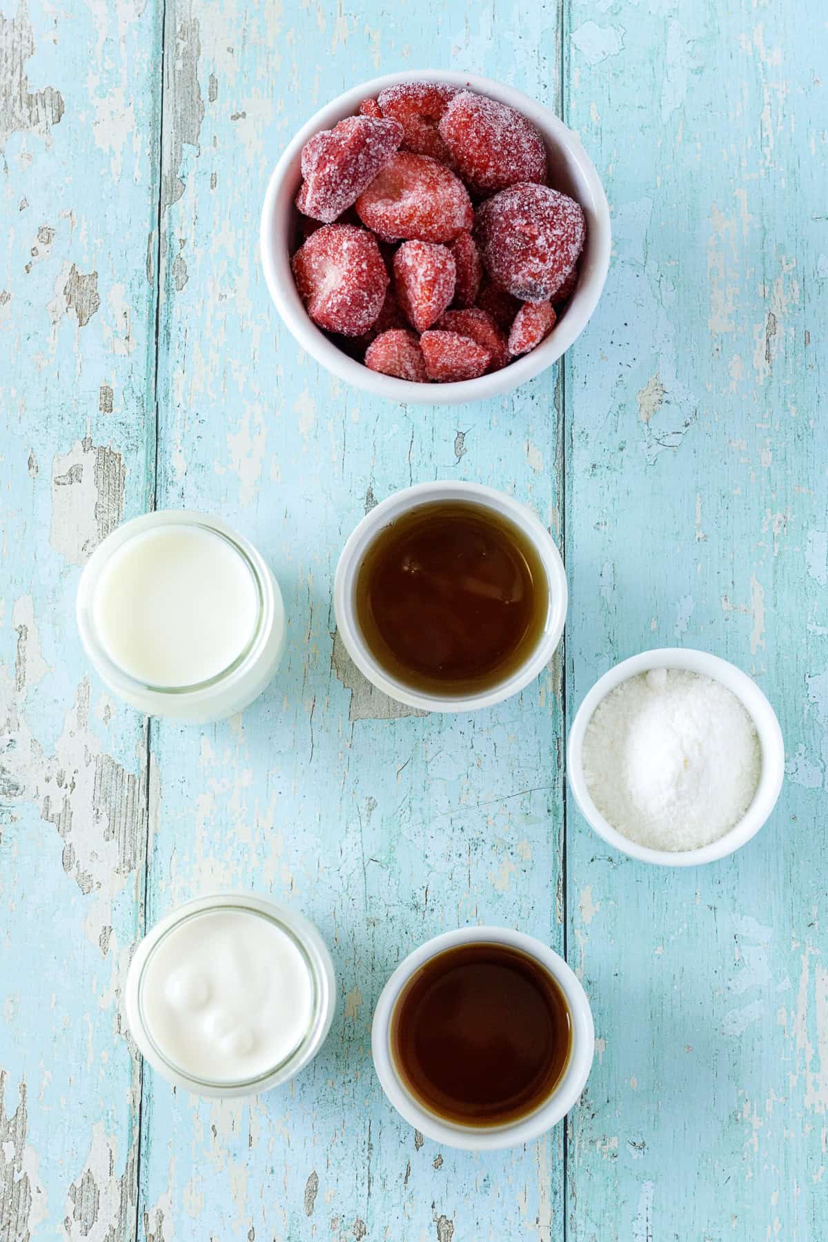 A bowl of frozen strawberries and small jars containing milk, maple syrup, vanilla extract, yogurt, and vanilla pudding are arranged on a distressed turquoise wooden surface.