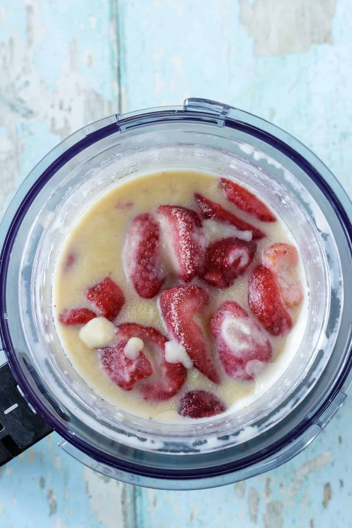 A blender filled with sliced strawberries and a creamy liquid mixture, ready to be blended, on a light blue wooden surface.