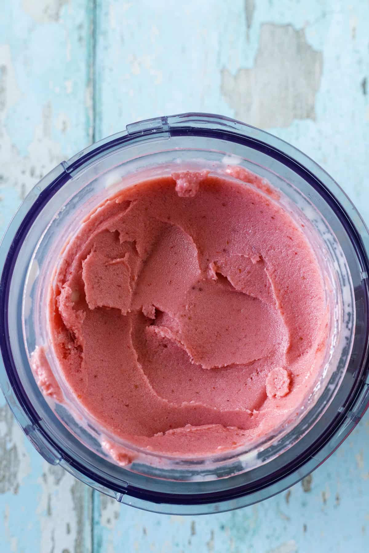 A blender container filled with smooth pink strawberry ice cream, viewed from above, on a distressed blue wooden surface.