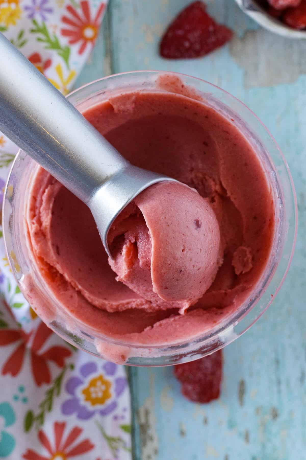 A metal ice cream scoop in a container of bright pink strawberry ice cream on a blue wooden surface, with a floral-patterned napkin nearby.