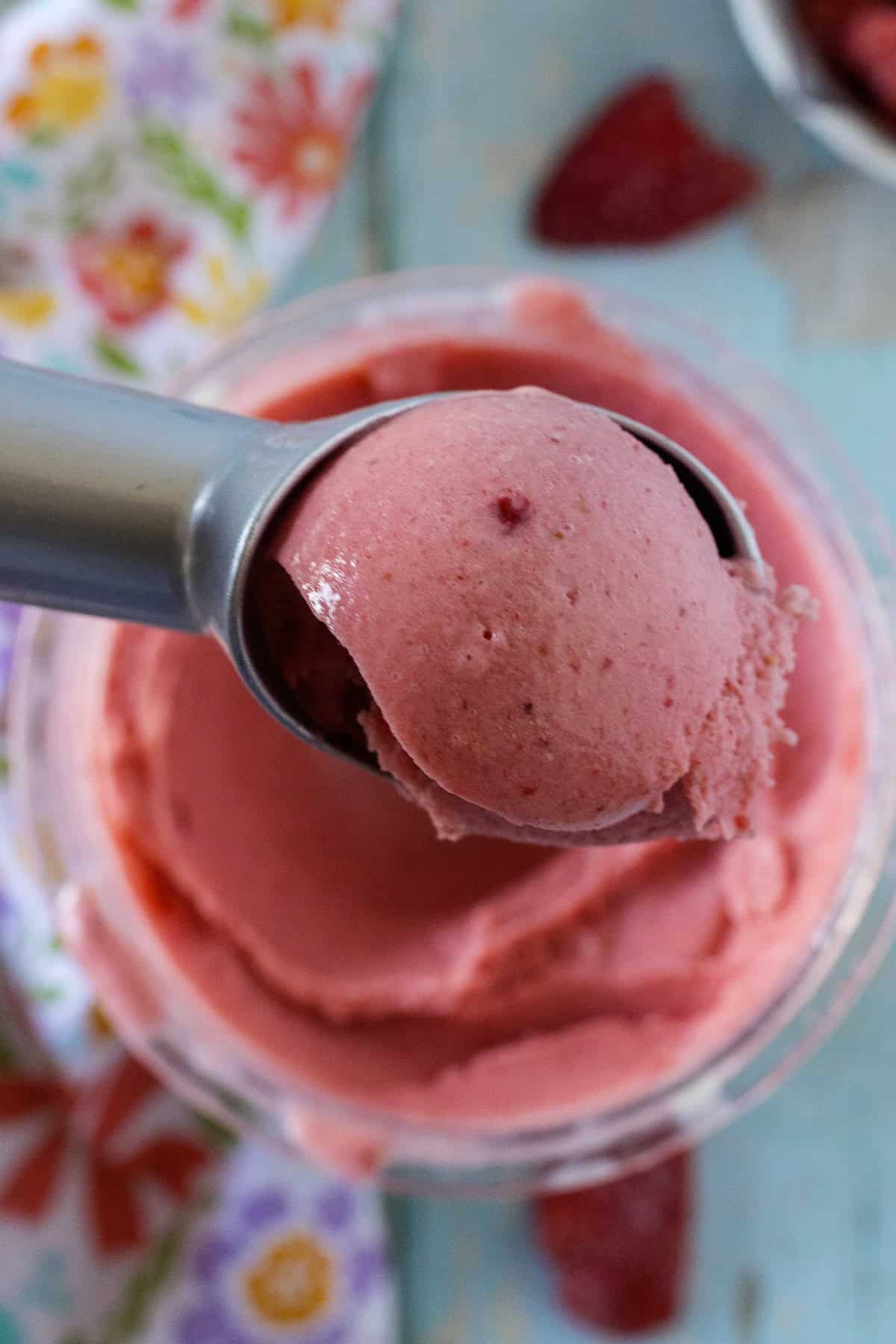 A metal scoop holds a serving of ninja creami strawberry ice cream above a container, with a floral cloth in the background.