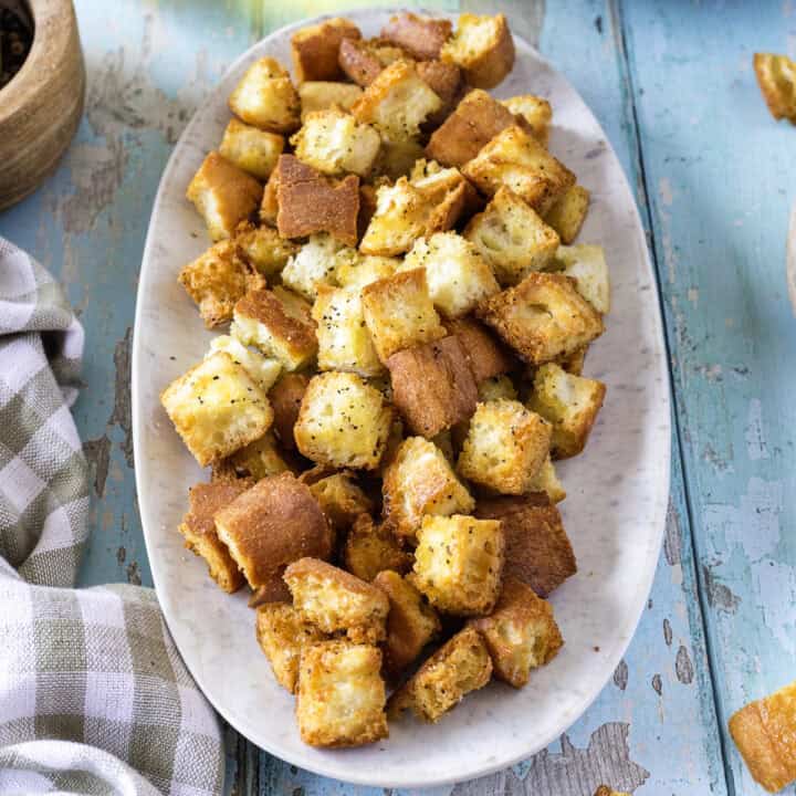 A white oval plate filled with golden-brown, seasoned air fryer croutons sits on a blue wooden table next to a checkered cloth.