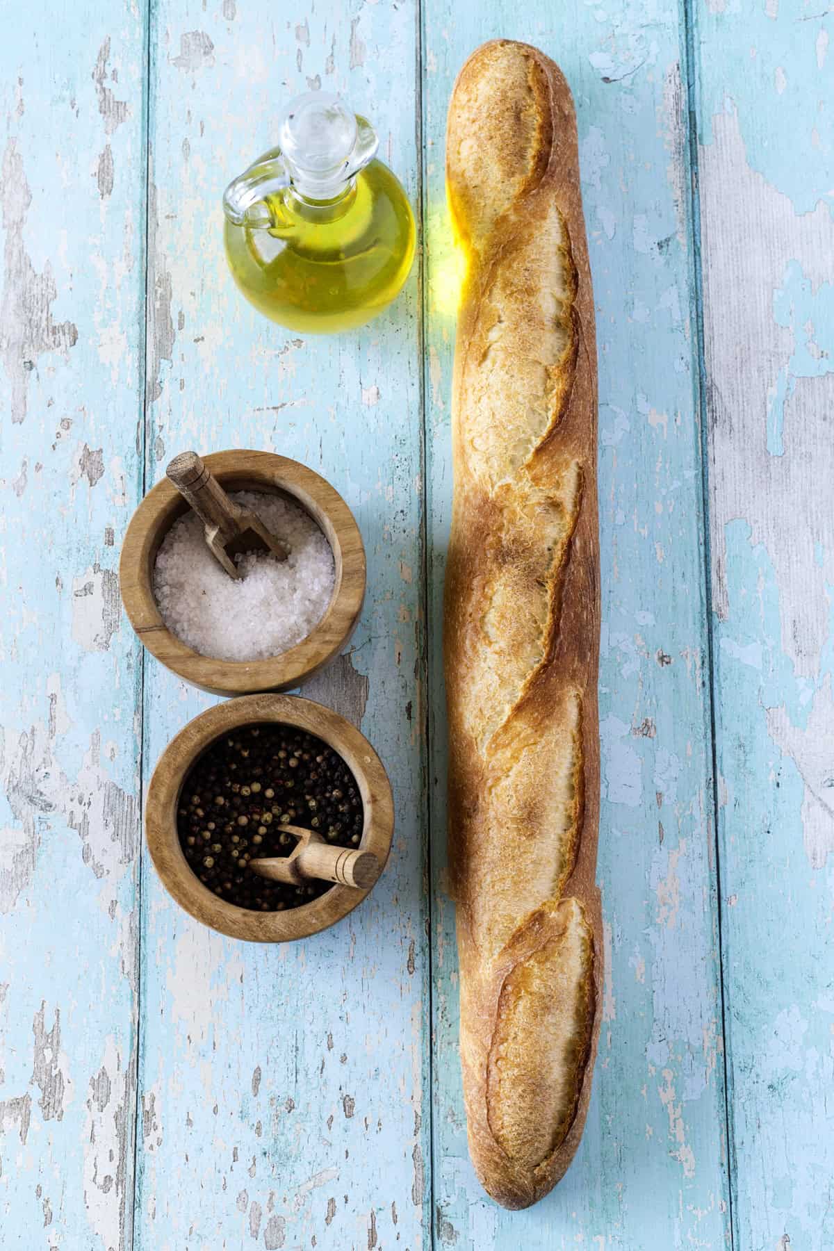 A baguette, a glass bottle of olive oil, a bowl of salt, and a bowl of mixed peppercorns are arranged on a rustic blue wooden surface.