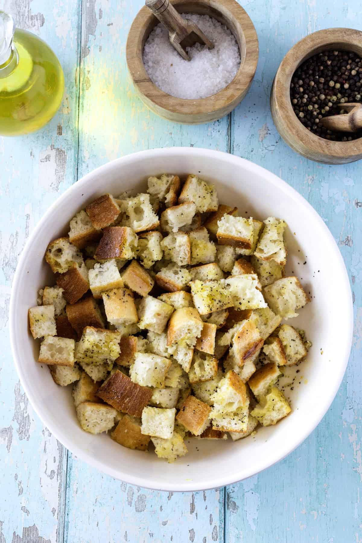 A white bowl filled with seasoned croutons is on a blue-painted wooden surface, next to a bottle of olive oil, a bowl of salt, and a bowl of pepper.