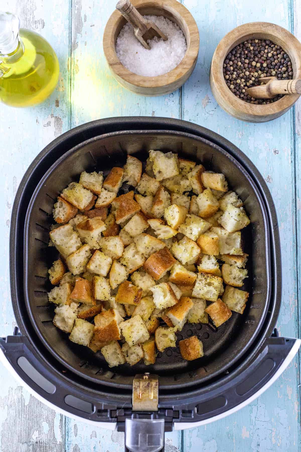 Cubed bread pieces in an air fryer basket, with oil, salt, and pepper containers on a blue wooden surface.