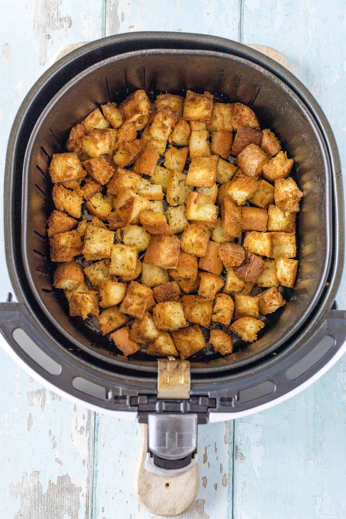 Crispy, golden-brown croutons inside an open air fryer basket on a light blue wooden surface.