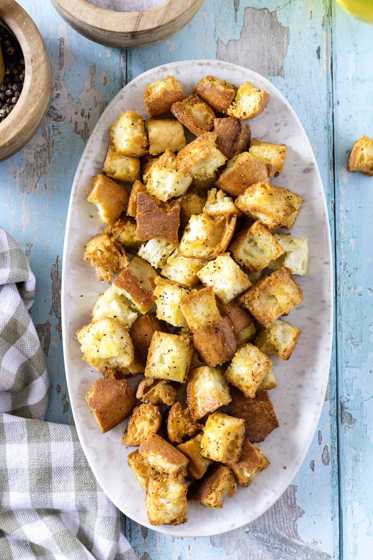 An oval plate filled with seasoned, toasted bread cubes sits on a rustic blue wooden surface.